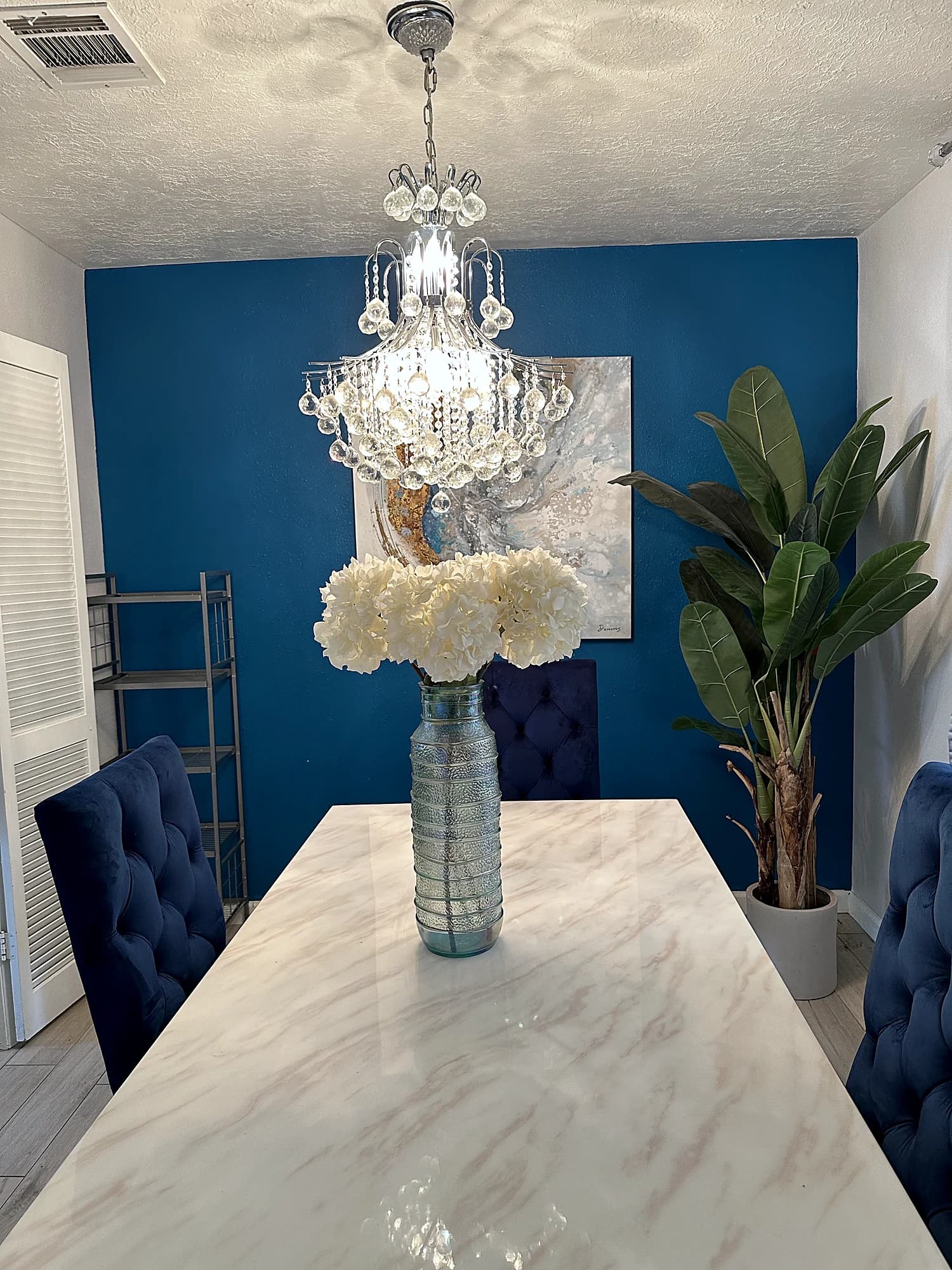 Dining room with a blue accent wall, a crystal chandelier, a marble look table and navy tufted chairs